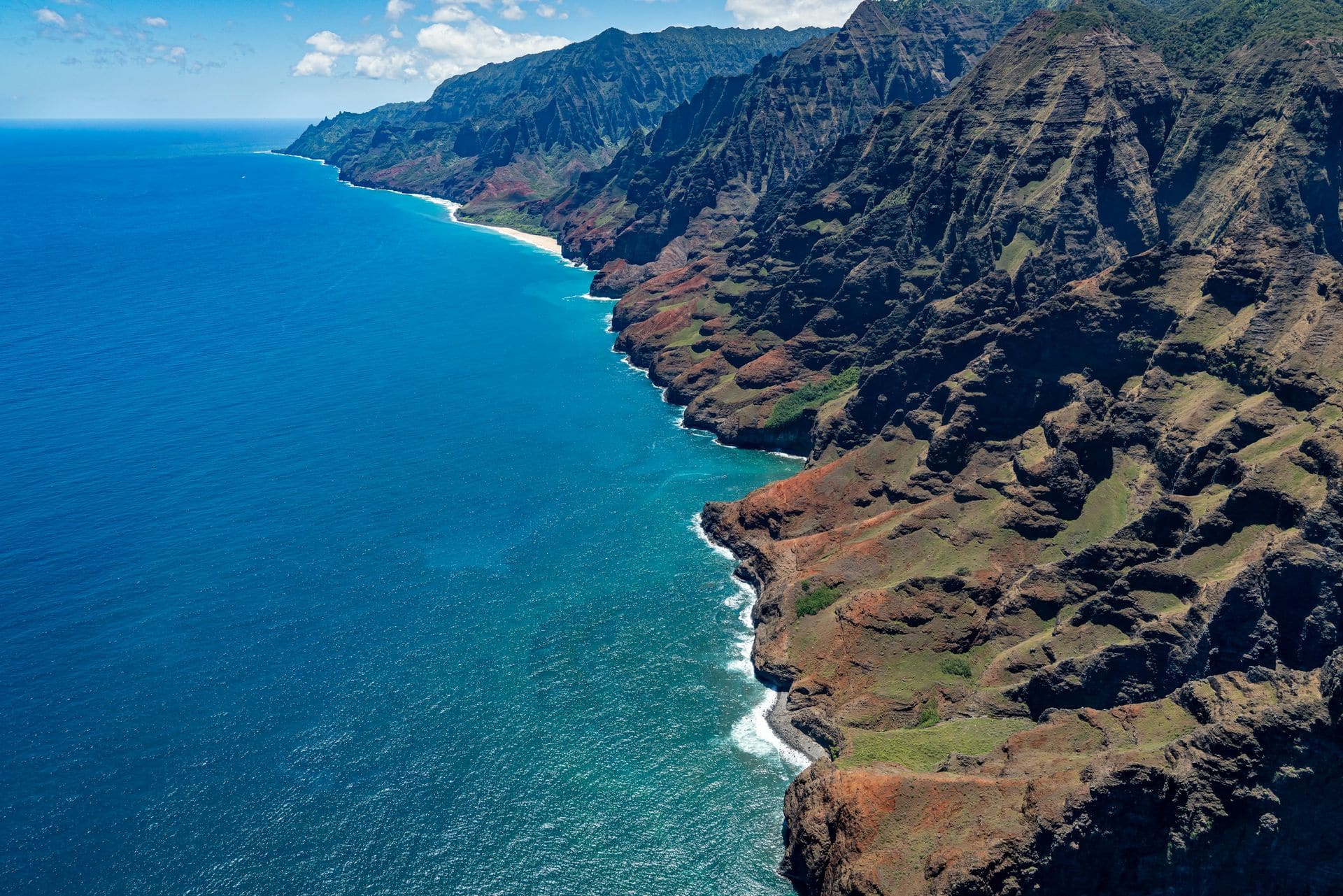 Aerial view of the Nā Pali Coast on Kauaʻi: fluted green and rust-red sea cliffs dropping into deep blue ocean