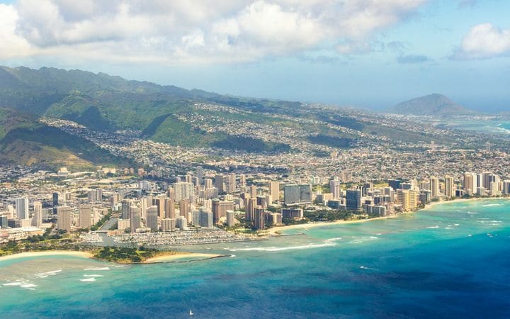 Aerial view of Honolulu’s high-rises along a turquoise shoreline, with green ridges behind