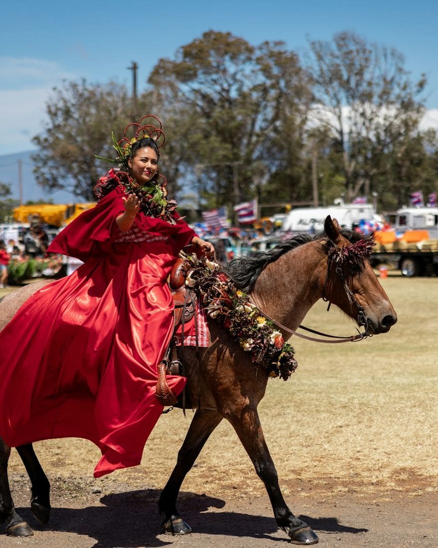 A pāʻū rider in a flowing red skirt and lei on horseback